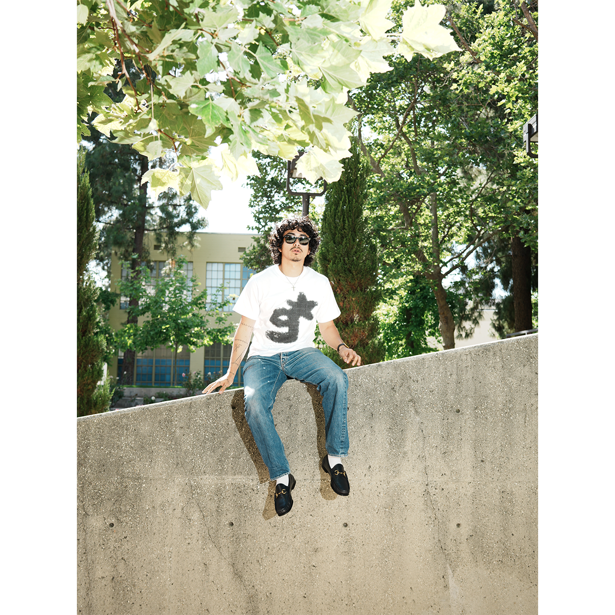Person sitting on a concrete ledge with trees in the background wearing g & star white tee
