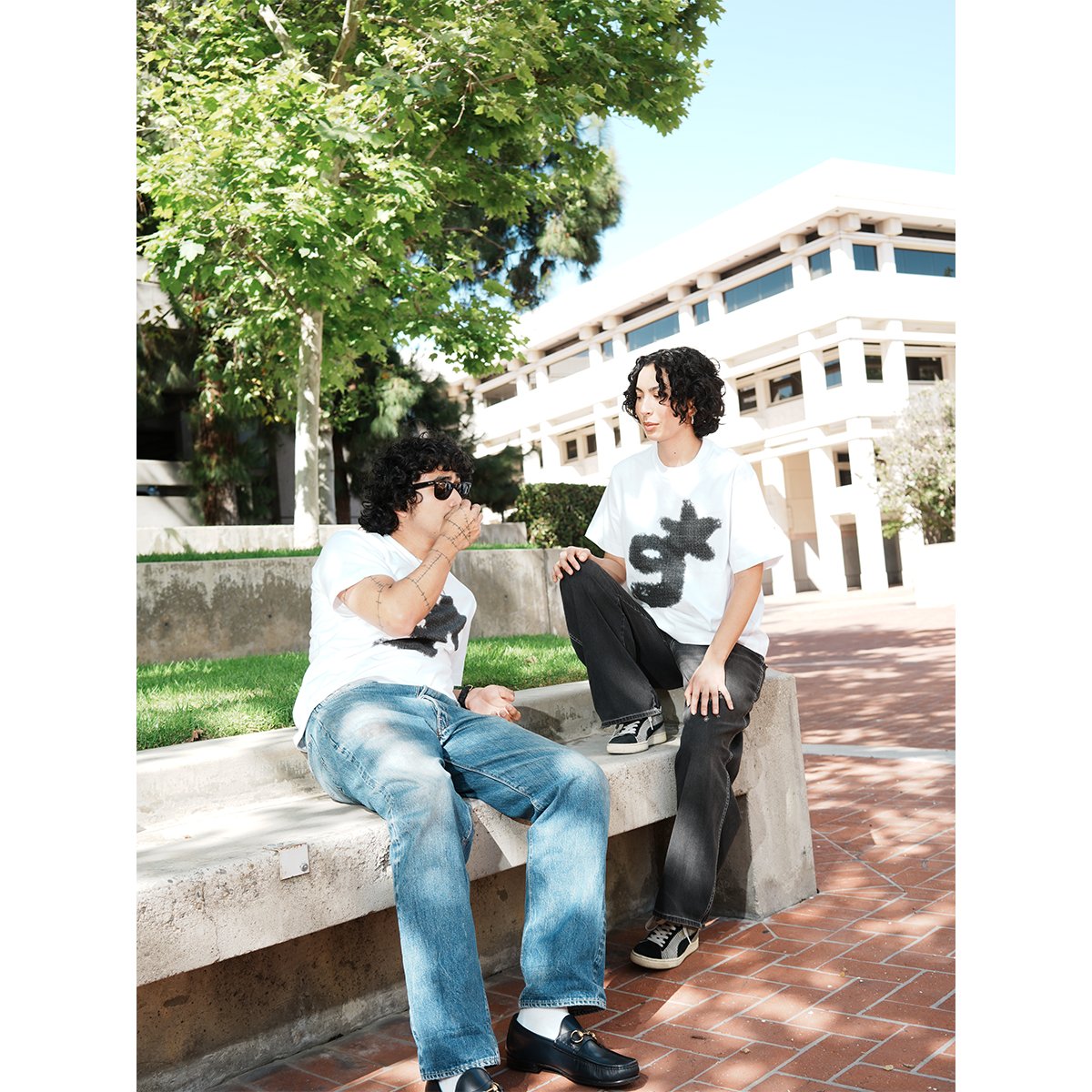 Two people sitting on a concrete bench outdoors with trees and a building in the background wearing g & star white tees